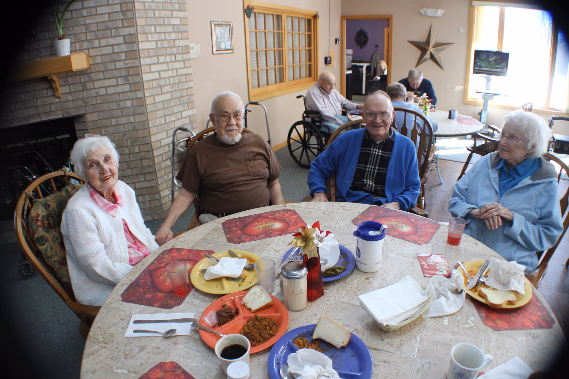 Several elderly residents seated around a table with plates, cups, and placemats in a senior living dining room.