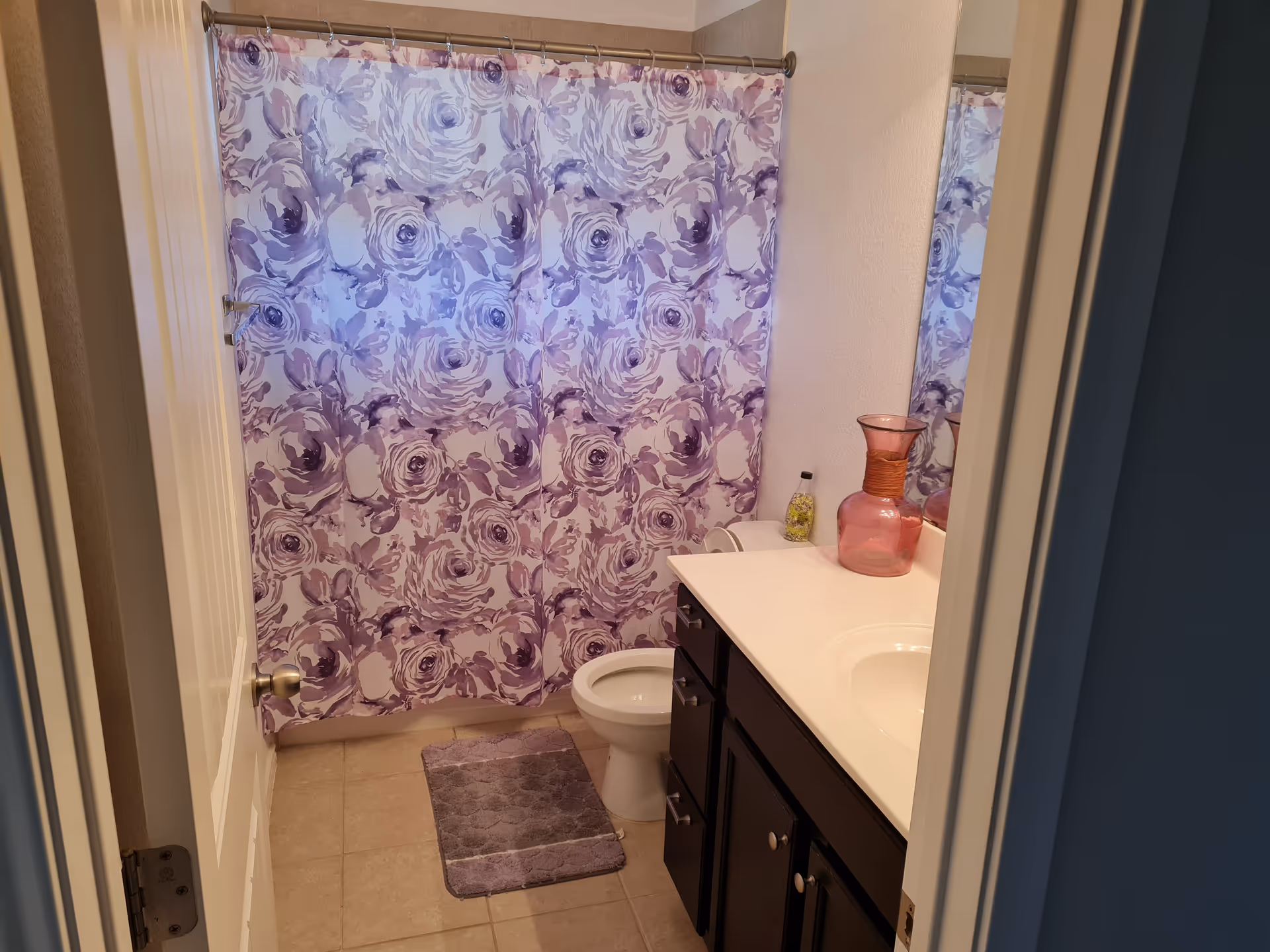 View into a bathroom with a white door partially open. Inside, there is a toilet, a dark wood vanity with a white countertop and sink, a large mirror above the sink, and a purple floral shower curtain covering the bathtub. A purple bath mat is placed on the tiled floor in front of the toilet. A pink decorative vase and a small bottle are on the countertop.