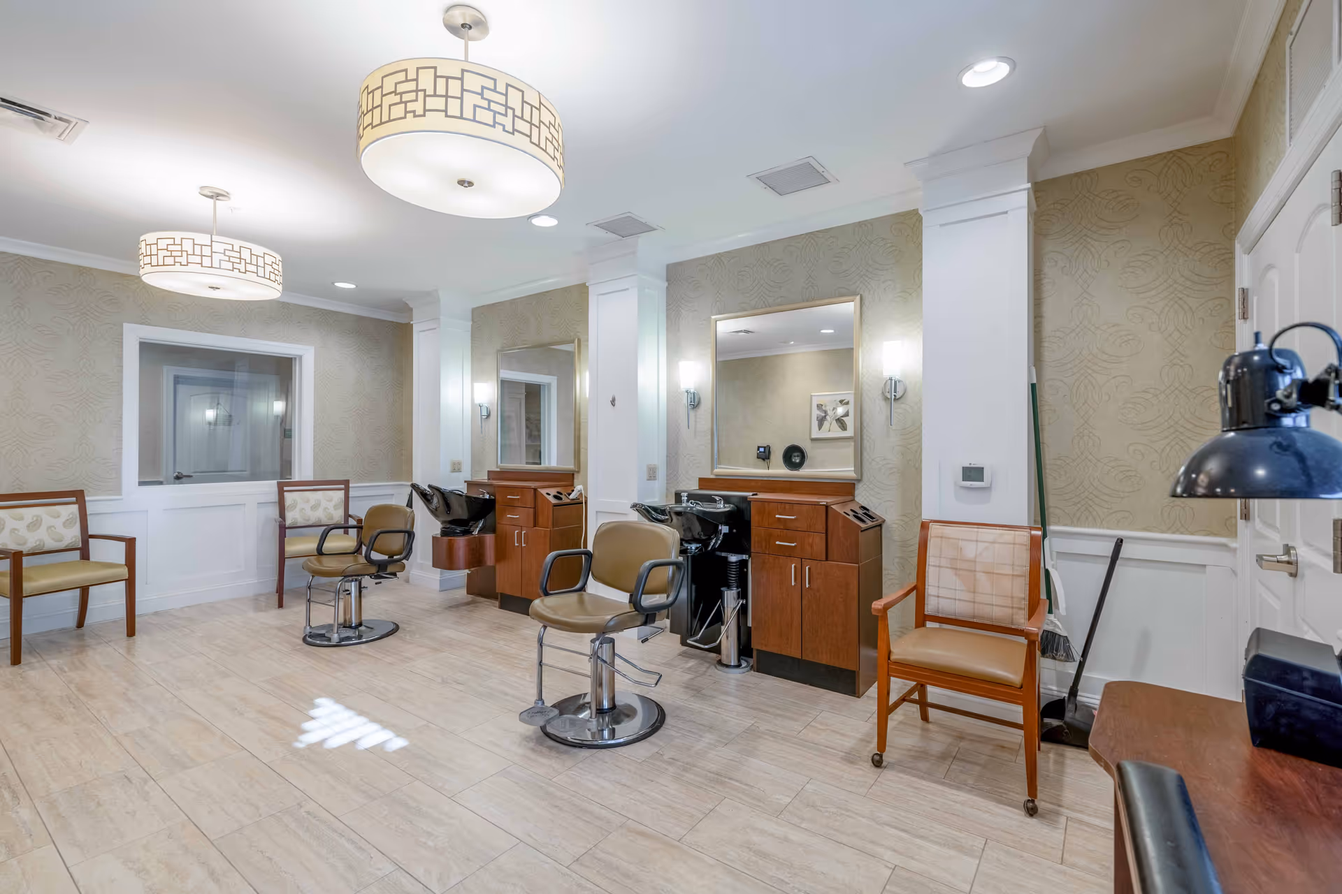 Interior view of a senior living facility's hair salon area featuring two salon chairs in front of mirrors and wash basins, with additional seating chairs along the walls, modern ceiling lights, and light-colored tiled flooring.