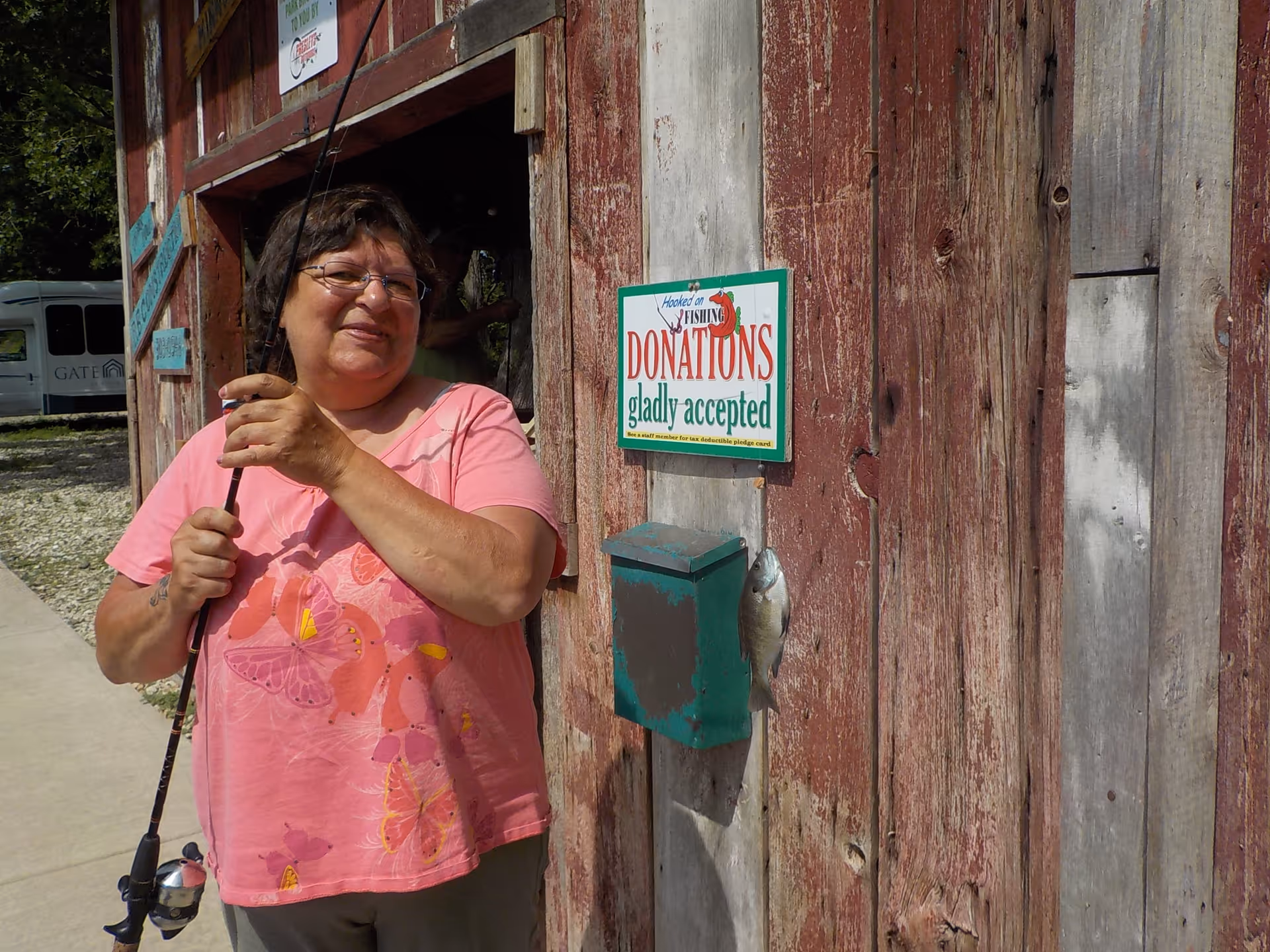 A woman wearing glasses and a pink shirt with butterfly designs is holding a fishing rod and standing next to a weathered wooden wall with a green donation box and a sign that reads 'Hooked on Fishing DONATIONS gladly accepted'. A small fish is hanging from the donation box. In the background, there is a white vehicle with the word 'GATE' visible on it.