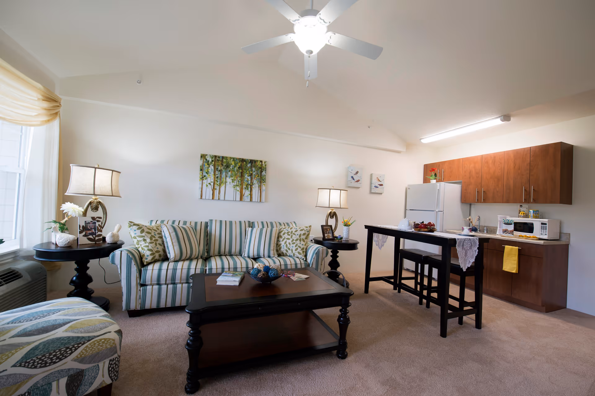 A cozy living area in a retirement facility featuring a striped sofa with patterned cushions, two side tables with lamps and decorative items, a coffee table with books and decor, and a small kitchen area with wooden cabinets, a refrigerator, microwave, and a high table with two stools. The room has beige carpeting, a ceiling fan with light, and wall art including a painting of trees and bird pictures.