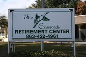 Outdoor sign for The Crossroads Retirement Center with a green bird logo and phone number 863-422-4961, situated on a lawn with bushes and trees in the background.