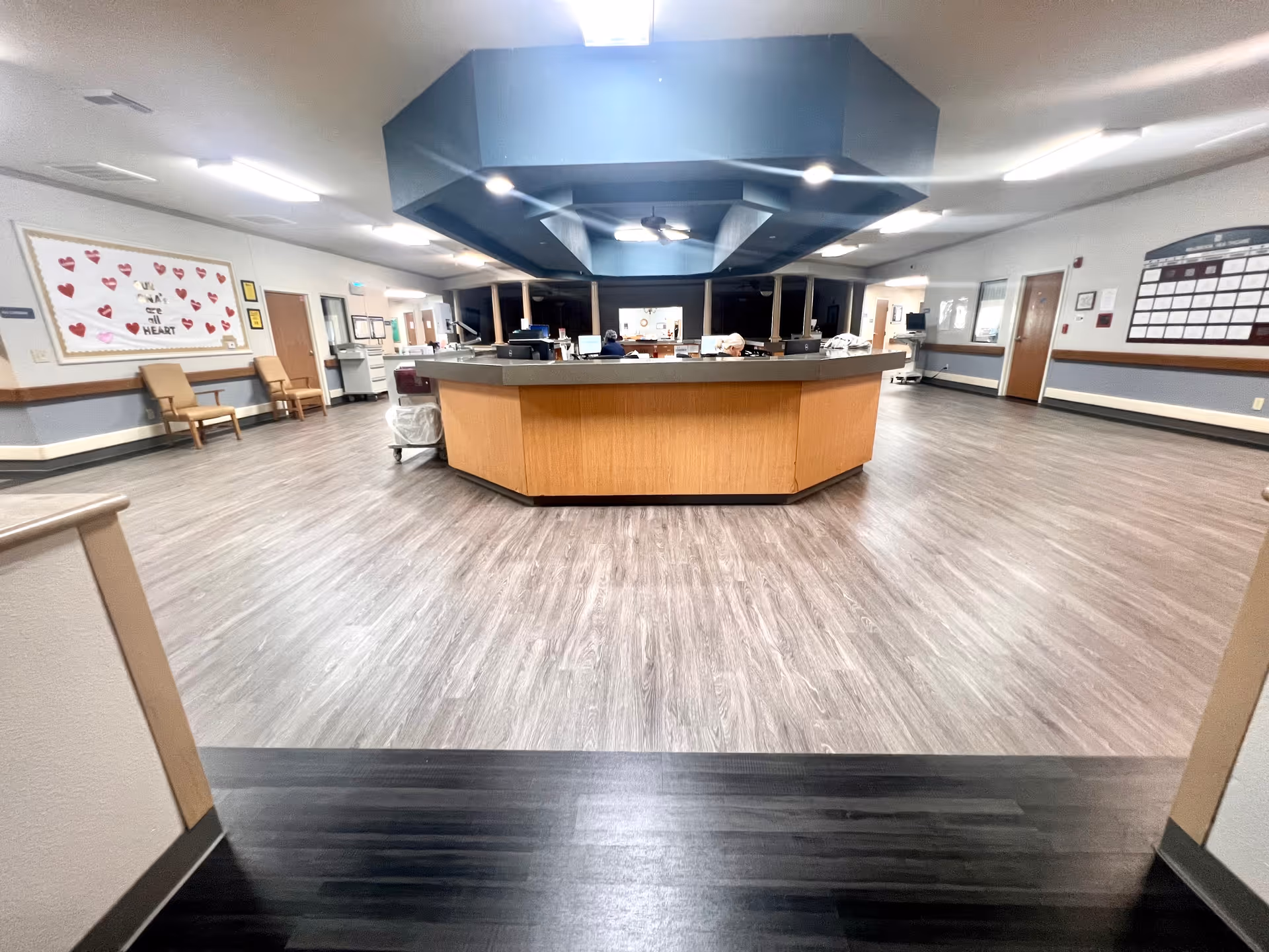 A wide view of a healthcare facility's reception or nurse station area with a large octagonal wooden desk in the center. The floor is covered with wood-patterned vinyl flooring, and there are chairs along the walls. Bulletin boards and informational displays are visible on the walls, and the lighting is bright with ceiling fixtures.