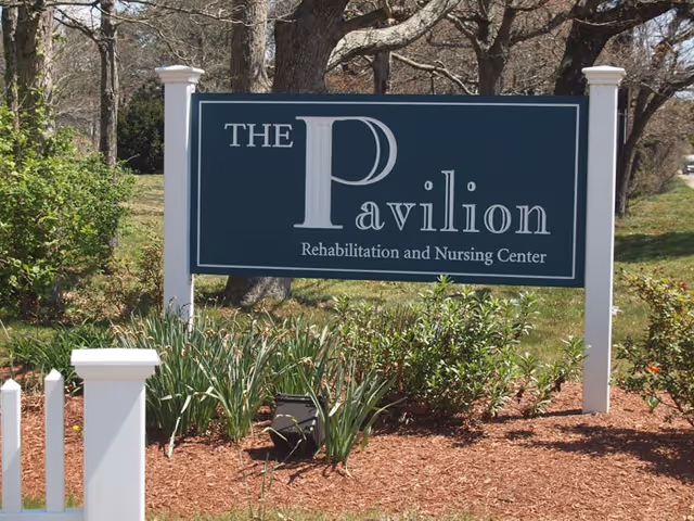 A large outdoor sign for The Pavilion Rehabilitation and Nursing Center surrounded by green bushes and plants with trees in the background.