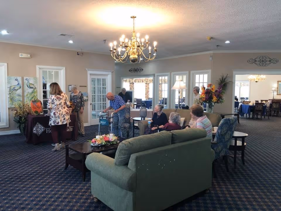 A common area in an assisted living community with several elderly residents sitting on sofas and chairs, and a few people standing near a table with decorations and artwork on the wall. The room has a chandelier, carpeted floor, and large windows and doors leading to another room with dining tables and chairs.