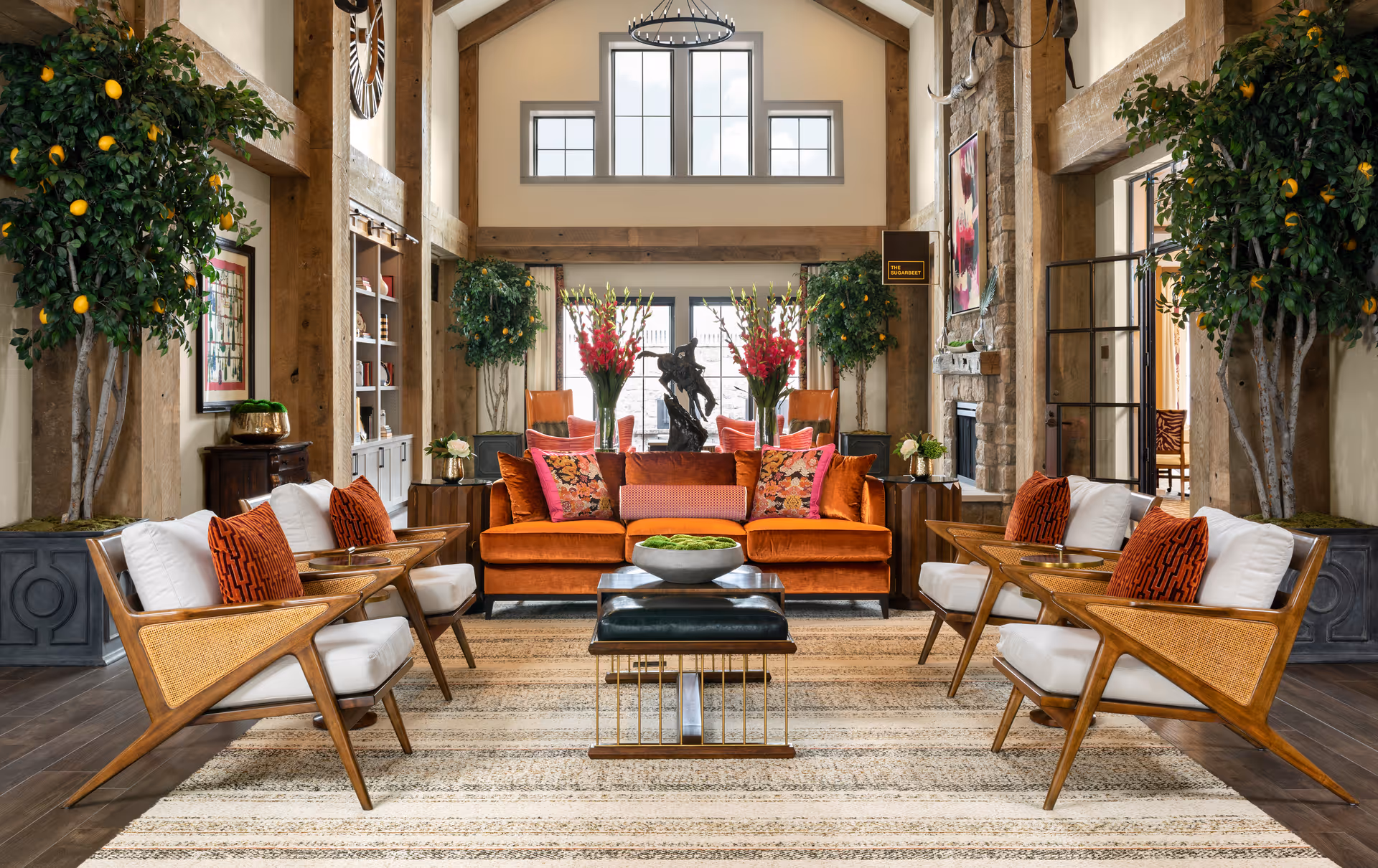 A bright, upscale lounge with a vaulted ceiling featuring an orange sofa, matching armchairs, coffee tables, potted trees, and a central rug.