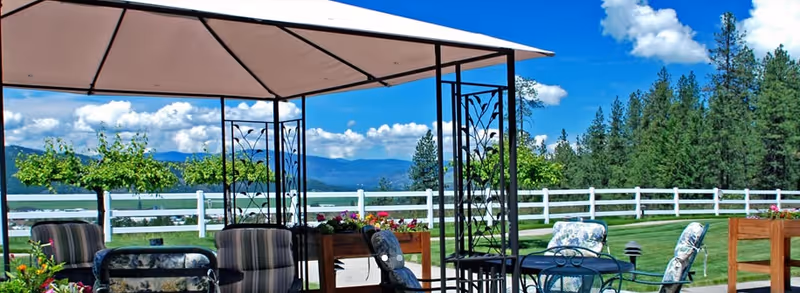 Outdoor patio area with a canopy providing shade over several metal chairs and tables. The patio overlooks a green lawn with flower beds and a white fence, with trees and mountains visible in the background under a blue sky with scattered clouds.