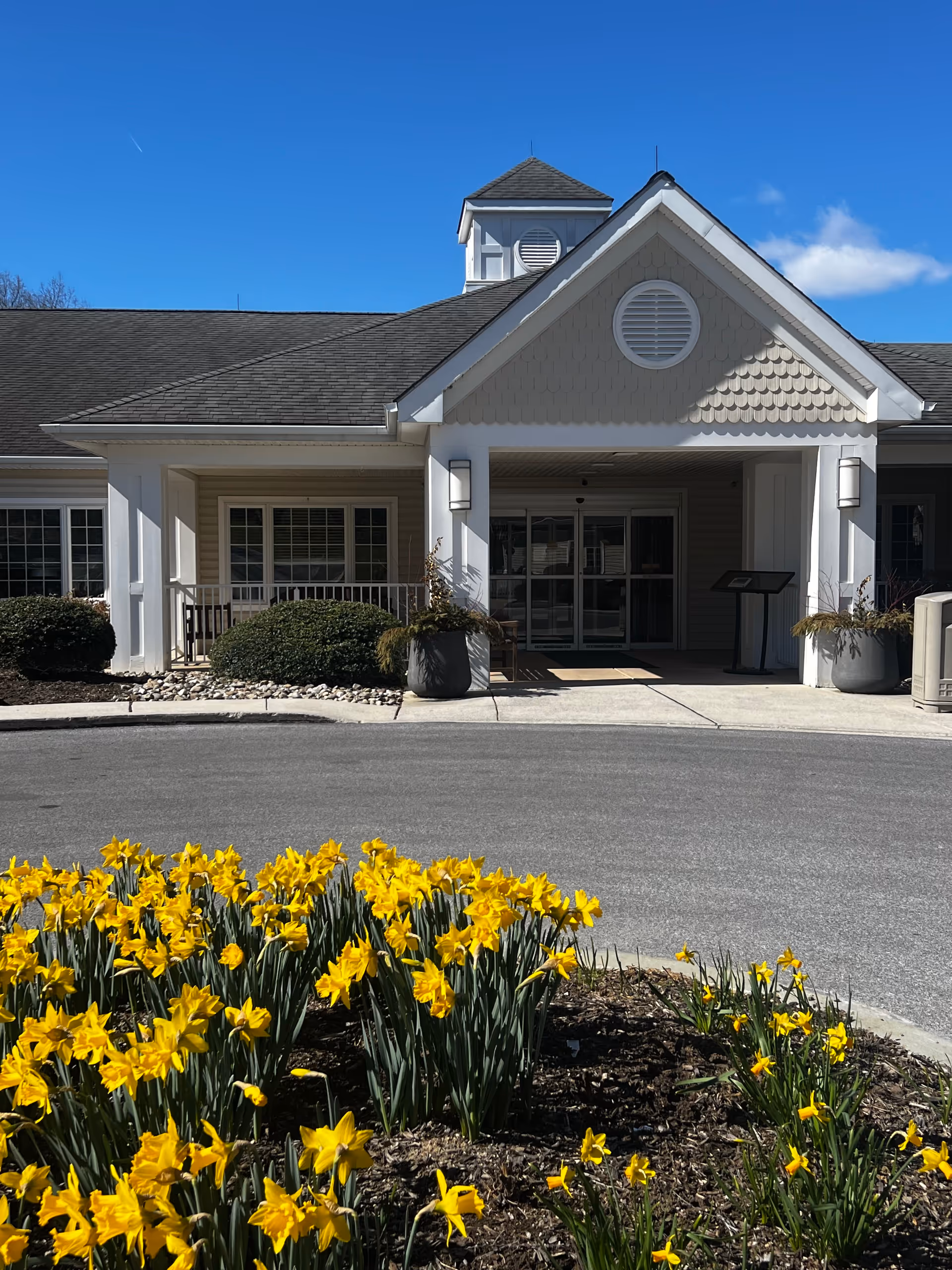 Entrance of a senior living facility named Barclay Friends, featuring a covered porch with white pillars, a peaked roof with decorative shingles, and large windows. In the foreground, there is a circular flower bed filled with blooming yellow daffodils under a clear blue sky.