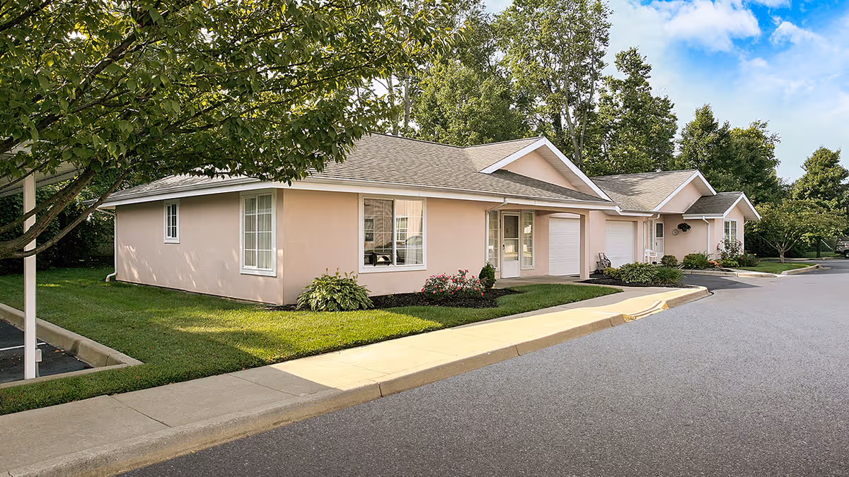 Exterior view of a single-story residential building with beige walls and a gray shingled roof, surrounded by green grass, trees, and a paved driveway under a partly cloudy blue sky.