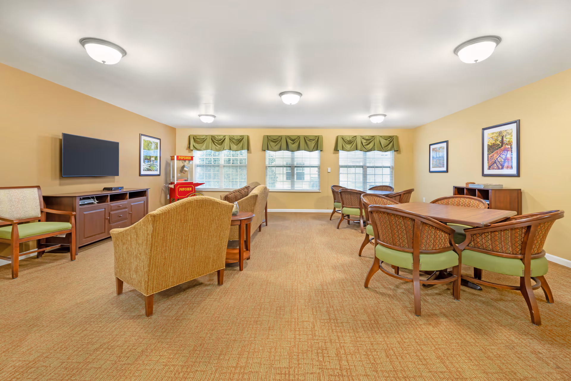 A well-lit common room with beige walls and carpet, featuring a flat-screen TV mounted on the wall, a popcorn machine, two sets of tables with green cushioned chairs, and two beige armchairs. The room has three windows with green valances and framed artwork on the walls.