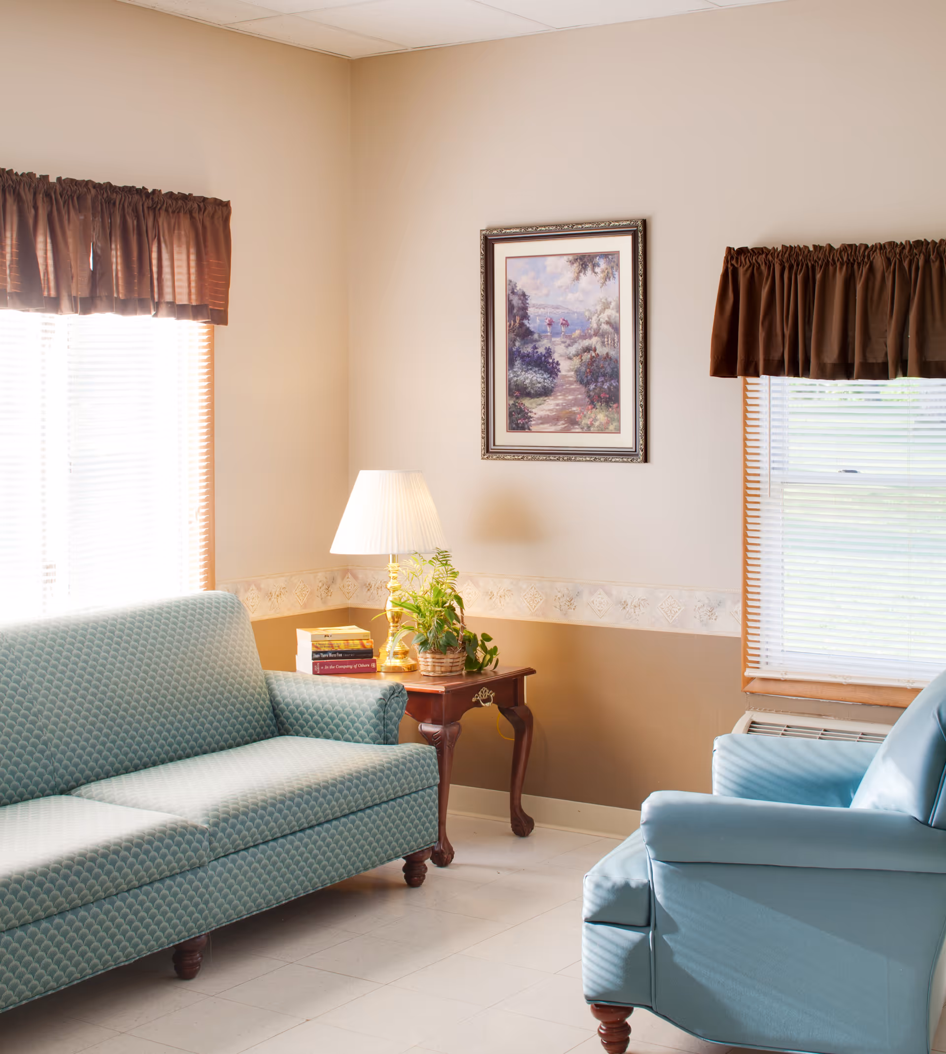 A cozy sitting area with teal sofa and armchair, a wooden side table with a lamp and plant, framed artwork on the wall and windows with brown valances.