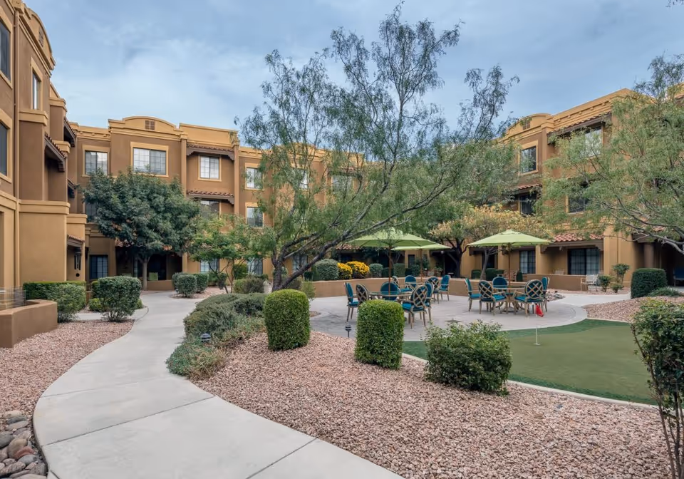 Outdoor courtyard area of a senior living facility with a curved concrete walkway, landscaped bushes and trees, several round tables with green umbrellas and chairs, and a small putting green with a red flag. The surrounding building is three stories tall with tan stucco walls and multiple windows.