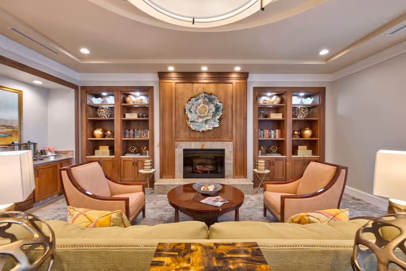 Cozy sitting area with a fireplace flanked by built-in wooden shelves, two armchairs, a coffee table and a sofa in a well-lit living room.