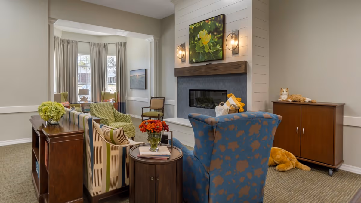 A cozy living room area with a blue patterned armchair, a striped sofa, and a green armchair arranged around a modern fireplace. The fireplace has a wooden mantel with a floral picture above it and two wall sconces on either side. There is a small round wooden table with a vase of red flowers and books, and a wooden cabinet with two stuffed animal cats on and near it. Large windows with beige curtains allow natural light into the room.