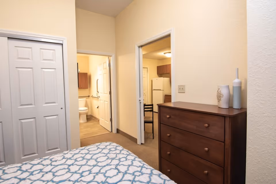 View from a bedroom showing a bed with a blue and white patterned bedspread, a wooden dresser with two decorative vases on top, a closet with white sliding doors, and two open doorways leading to a bathroom with a toilet and a kitchen area with a refrigerator and cabinets.