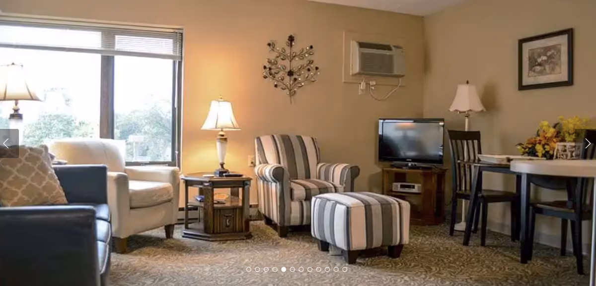 Comfortable common living area with upholstered chairs, a striped ottoman, side tables, a TV and a small dining table.