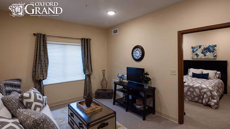 A cozy living room with a white couch adorned with patterned pillows, a window with patterned curtains, a coffee table with decorative items, and a TV on a black stand. Through an open doorway, a bedroom is visible with a bed covered in a geometric patterned comforter and a world map artwork on the wall above the headboard.