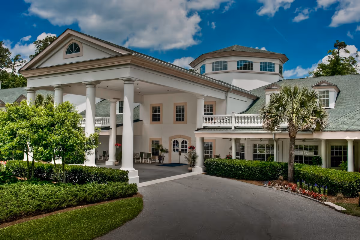 Front exterior view of a large white building with classical columns and a covered entrance driveway. The building is surrounded by well-maintained greenery, including bushes, trees, and a palm tree. The sky is partly cloudy with blue patches visible.