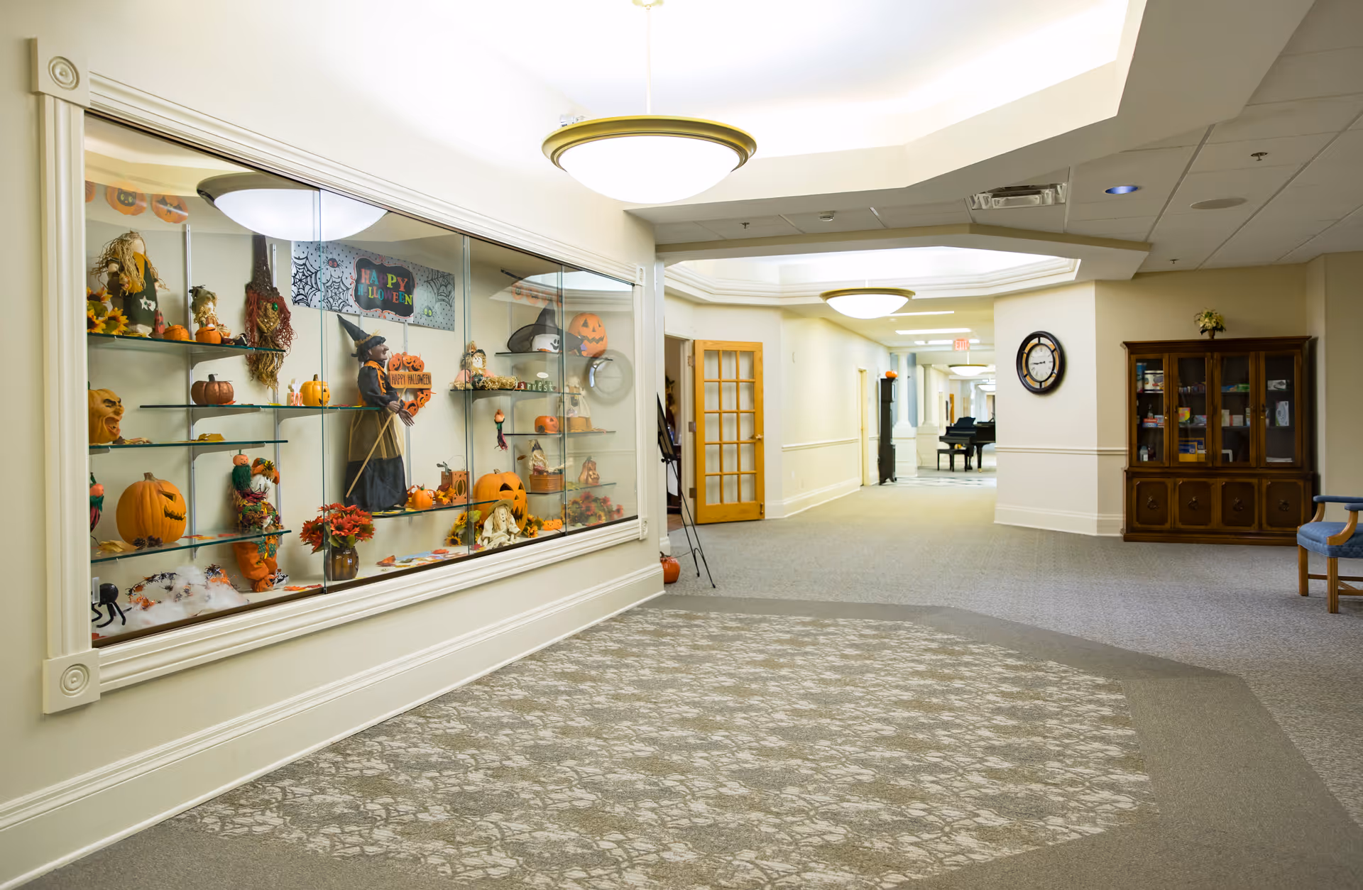 A well-lit hallway in a senior living facility decorated for Halloween with a glass display case filled with pumpkins, Halloween figurines, and seasonal decorations. The hallway has patterned carpeting, a wooden cabinet with glass doors, a wall clock, and a piano visible in the distance.