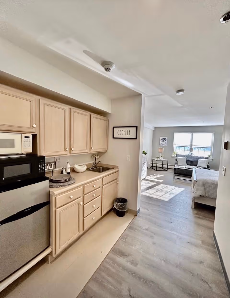 Interior view of a senior living facility unit showing a small kitchen area with light wood cabinets, a microwave, mini refrigerator, and a sink. Beyond the kitchen is a living and sleeping area with a bed, a couch, a coffee table, and a large window letting in natural light. A framed sign on the wall reads 'COFFEE.'