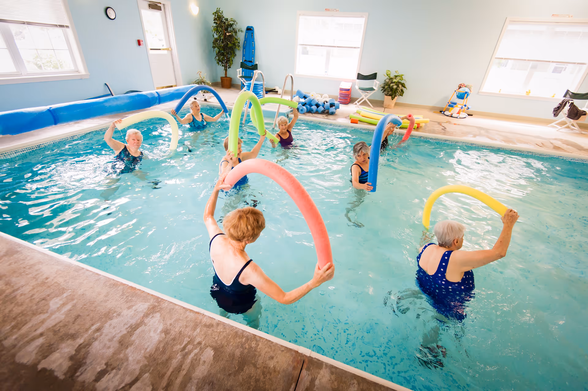 A group of elderly women participating in a water exercise class in an indoor swimming pool, each holding colorful pool noodles. The room has light blue walls, large windows, and pool equipment stacked in the background.