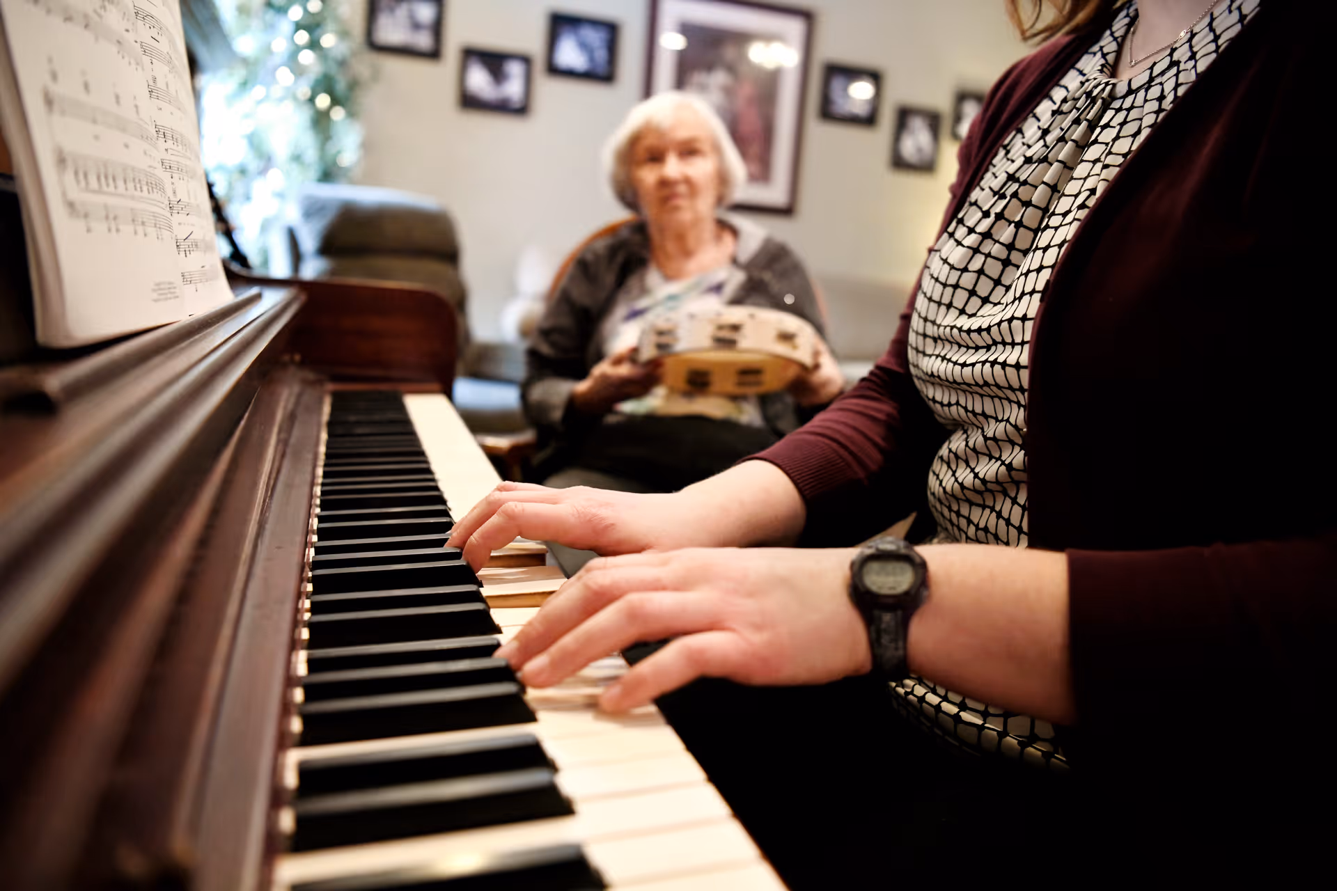 A person's hands play a piano in the foreground while an elderly woman holding a tambourine sits blurred in a cozy living room background.