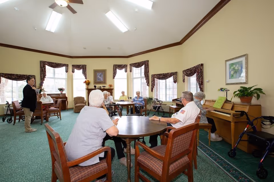 A group of elderly people sitting around tables in a spacious, well-lit common room with large windows, green carpet, and beige walls. A woman stands speaking to the group, and there is a piano with a plant on top in the corner. Several walkers are visible near the chairs.