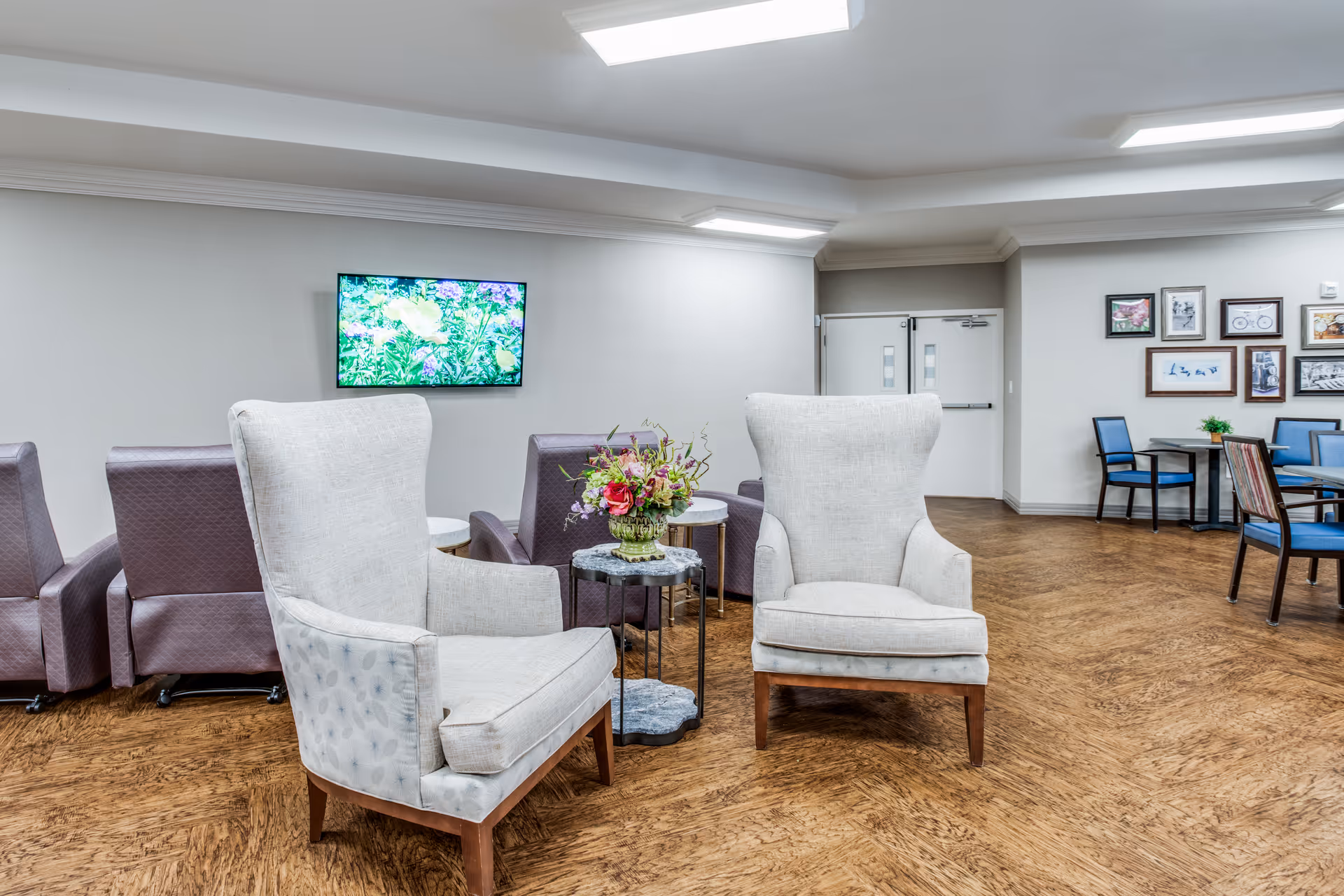 Bright senior living common room with two upholstered armchairs around a small table with flowers, extra seating, a wall-mounted TV, and dining tables in the background.