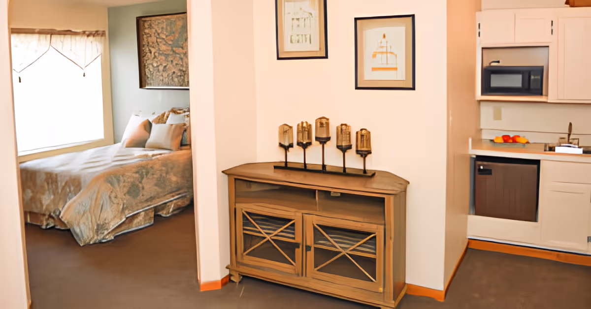Interior view of a senior living suite showing a bedroom through an open doorway, a wooden sideboard against a wall, and a small kitchenette to the right.