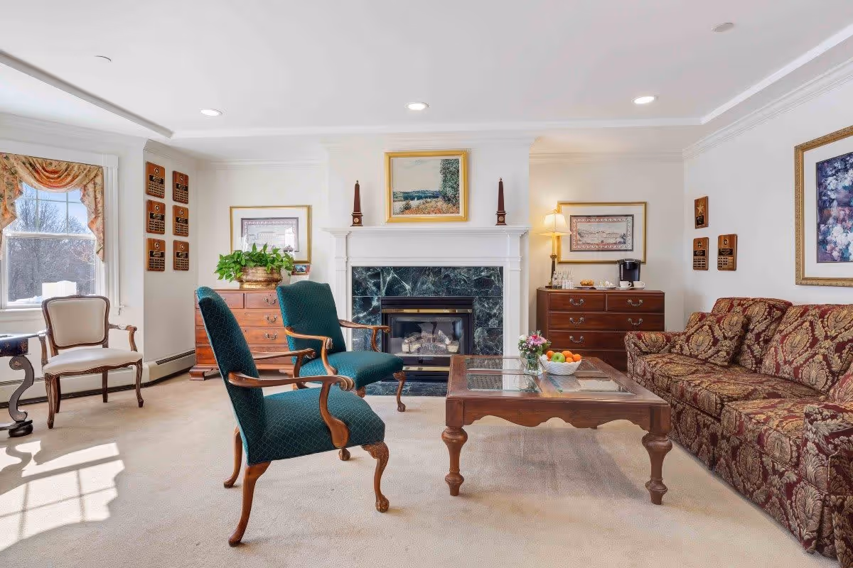 A bright and elegant living room with a fireplace centered on the far wall, flanked by two wooden dressers with framed artwork above them. Two green upholstered armchairs and a patterned sofa surround a wooden coffee table with a bowl of fruit and a small flower arrangement. A window with draped curtains lets in natural light, and the room has a beige carpet and white walls.