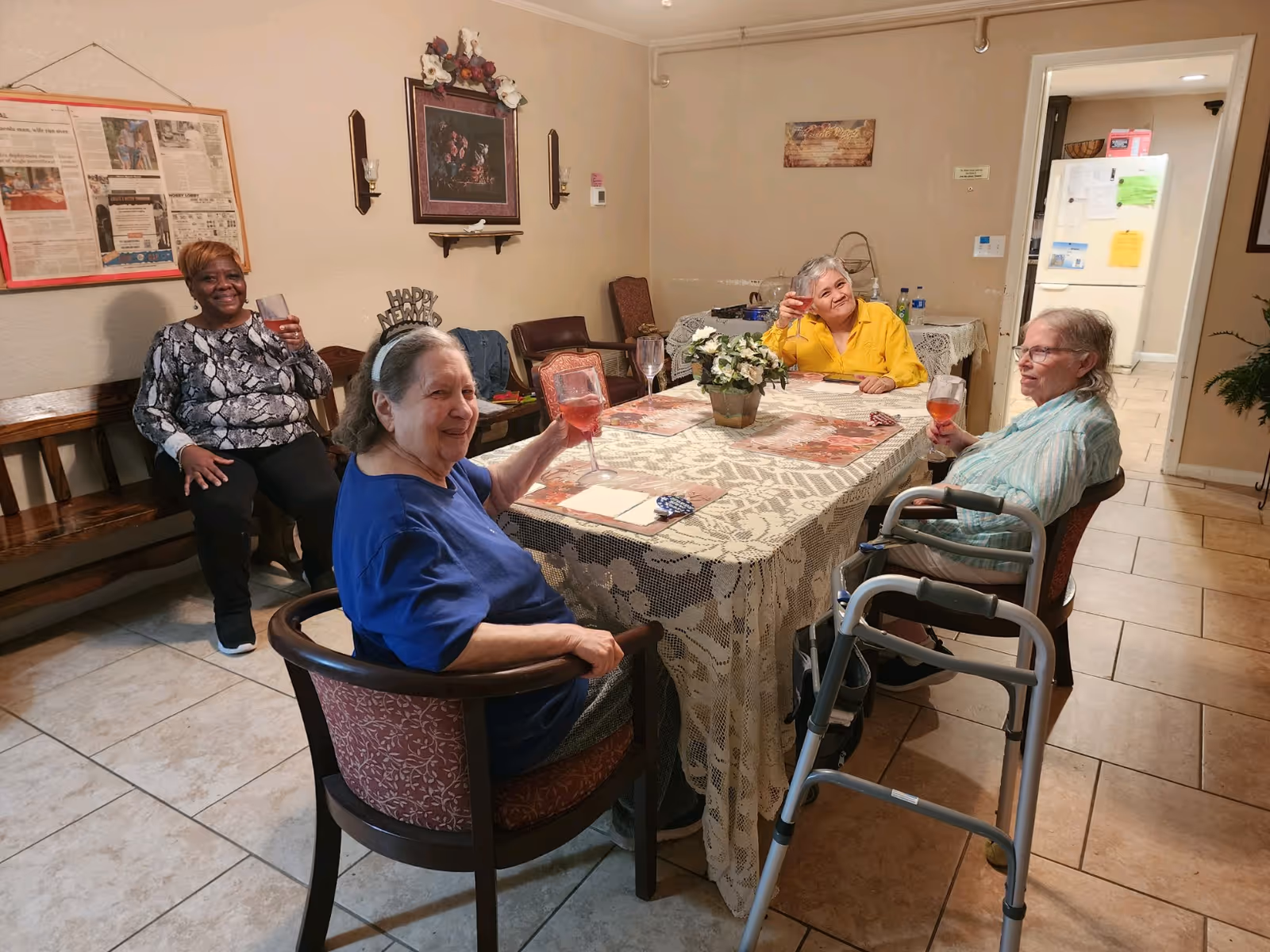 Four elderly women sitting around a dining table covered with a lace tablecloth, holding glasses with a pink beverage, smiling and enjoying each other's company in a cozy room. One woman is seated on a bench against the wall, and another woman has a walker next to her chair. The room has tiled floors, framed pictures on the walls, and a doorway leading to a kitchen area with a refrigerator.