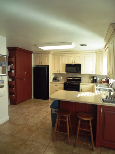 A kitchen with cream-colored upper cabinets and red lower cabinets, a black refrigerator, black microwave, and black stove. There is a beige countertop with two wooden stools underneath, a double sink, and various kitchen appliances on the counter. The floor is tiled in a light brown color.