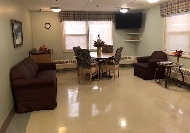 A cozy common area in Sheriden Woods Health Care Center featuring a brown sofa on the left, a round table with four floral-patterned chairs in the center, and a single armchair with a small side table holding a flower arrangement on the right. The room has three windows with plaid valances, a wall-mounted TV in the corner, a clock on the wall, and a painting above the sofa.