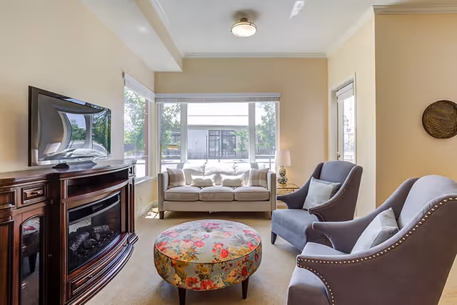 A cozy living room with a floral round ottoman in the center, two gray armchairs with light-colored pillows, a beige sofa by large windows letting in natural light, a wooden cabinet with an electric fireplace and a flat-screen TV mounted above it, and a small side table with a lamp.