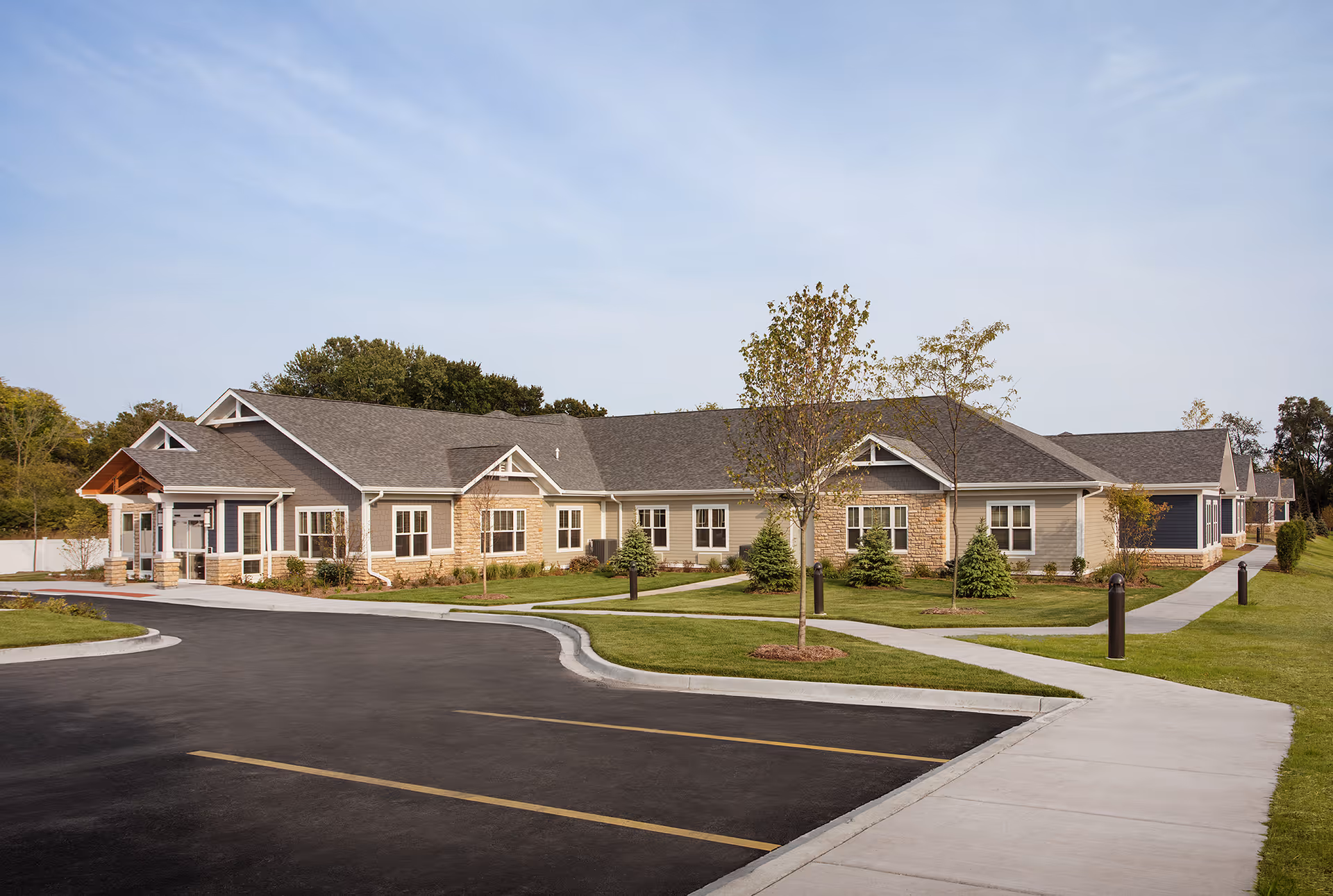 Exterior view of a single-story senior living facility building with a combination of stone and siding on the facade, surrounded by a well-maintained lawn, young trees, and paved walkways under a clear sky.