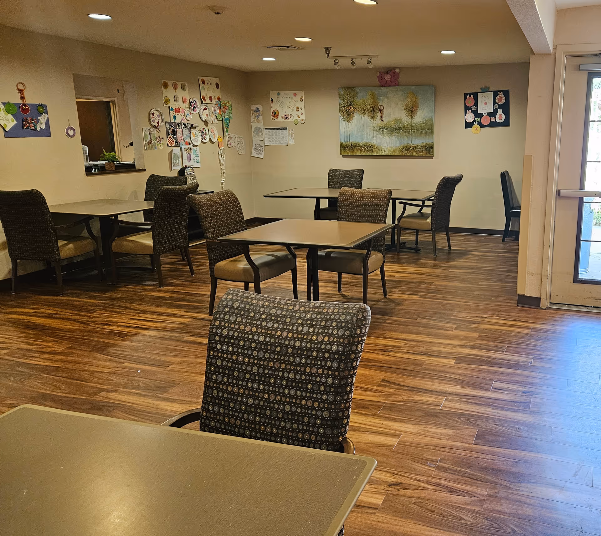 Interior view of a senior living facility dining room with several tables and cushioned chairs arranged on a wooden floor. The walls are decorated with colorful artwork and drawings, and there is a large painting of trees on one wall. A glass door is visible on the right side, allowing natural light into the room.