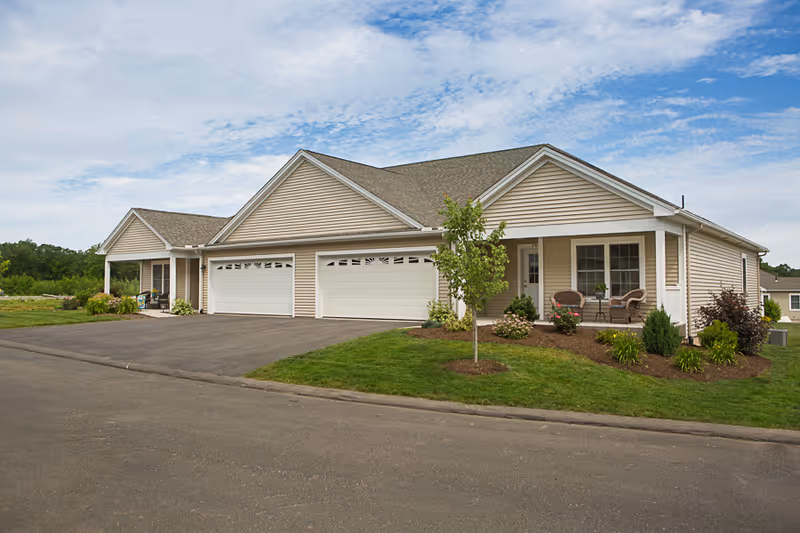 Exterior view of a beige single-story residential building with two garages and a small front porch with chairs and a table. The building is surrounded by a well-maintained lawn and landscaping, under a partly cloudy sky.