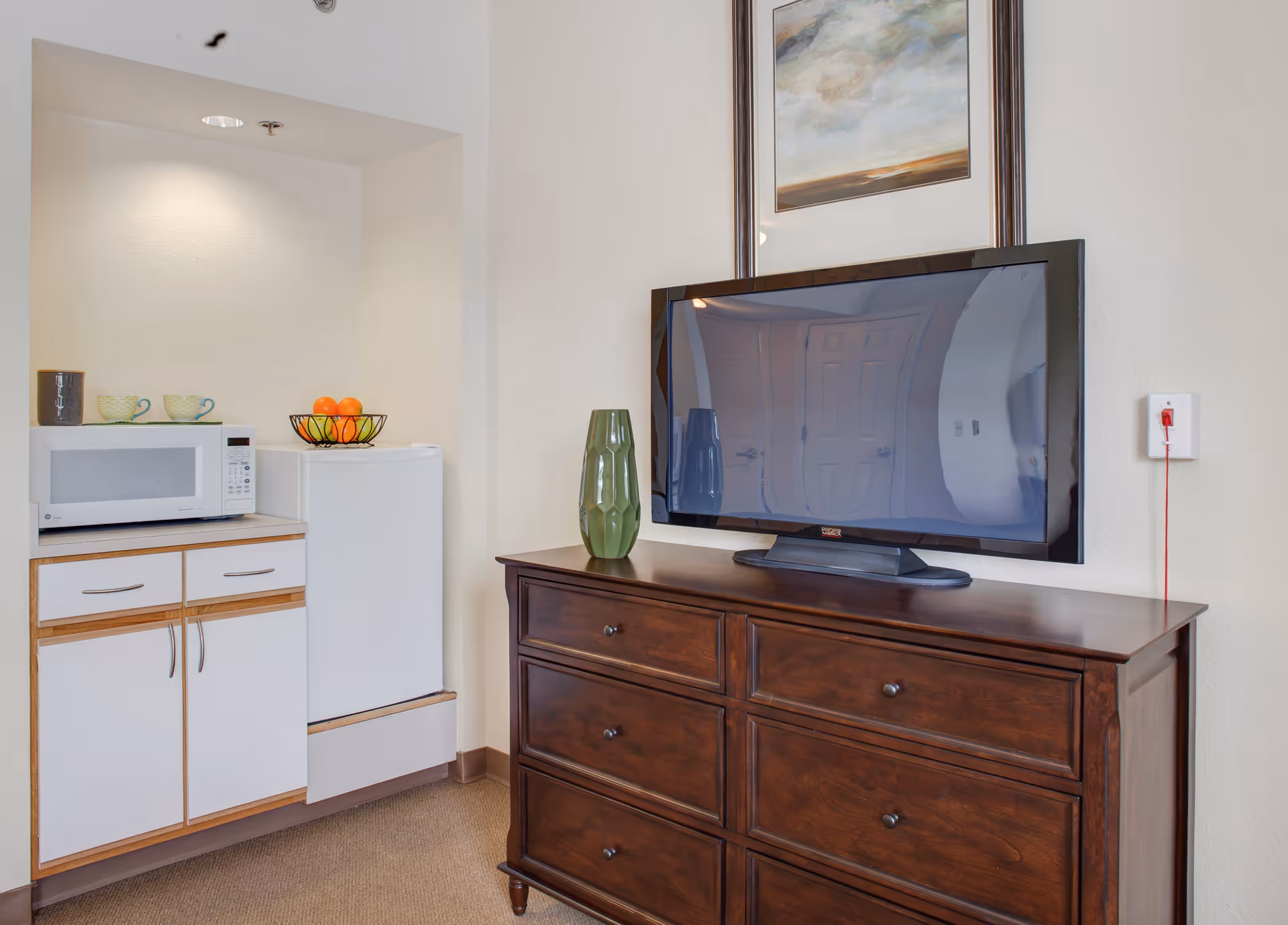 A small kitchenette area with a white microwave on a countertop, a mini refrigerator, and a fruit basket with oranges. Next to the kitchenette is a dark wooden dresser with six drawers, on top of which sits a flat-screen TV and a green decorative vase. Above the TV is a framed painting with abstract clouds and landscape.