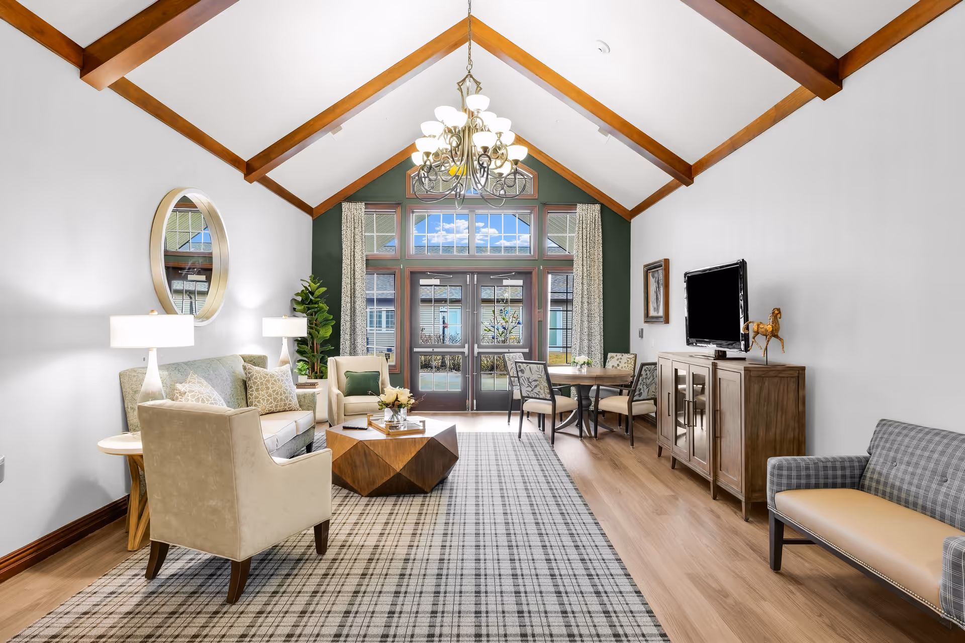 Bright communal living room with a vaulted beamed ceiling, seating area, TV cabinet and dining table facing large glass doors.