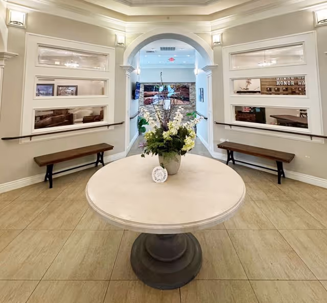 Interior entryway of an assisted living facility with a round table holding a floral centerpiece, benches along the walls, and an arched doorway leading to a seating area.