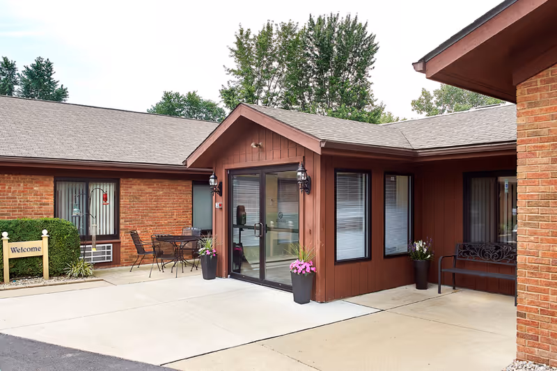 Front entrance courtyard of a brick and wood senior living building with glass double doors, potted flowers, a welcome sign and outdoor seating.