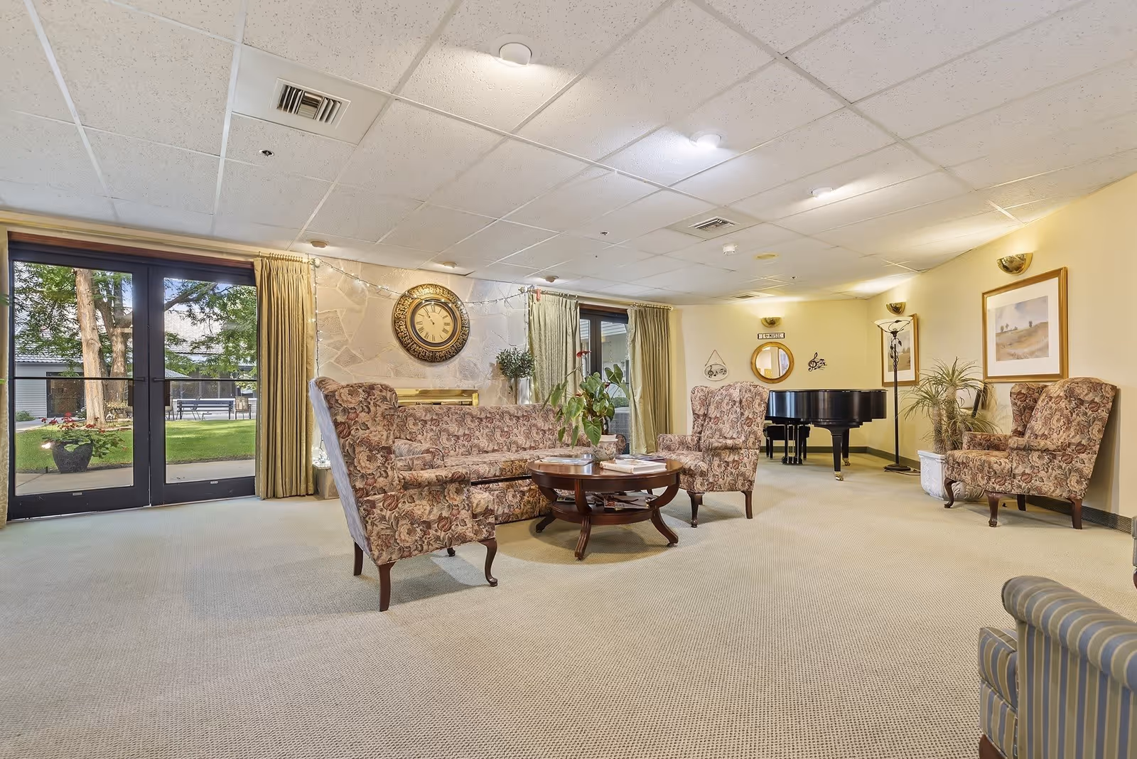 A senior living facility common area with floral upholstered armchairs and a matching sofa arranged around a wooden coffee table. The room has beige carpet, light yellow walls, and a ceiling with recessed lighting. A large decorative clock hangs on a stone accent wall, and a black grand piano is visible in the background. Glass doors with curtains lead to an outdoor garden area with trees and benches.