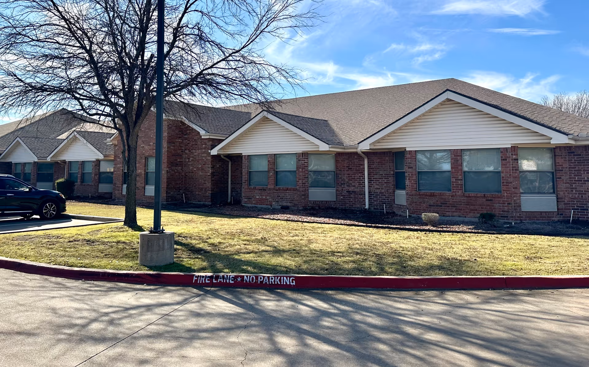 Exterior view of a single-story brick building with multiple windows and a sloped roof under a blue sky. There is a leafless tree and a lamp post on a grassy area next to a parking lot with a black car parked. The curb is painted red with white text reading 'FIRE LANE NO PARKING'.