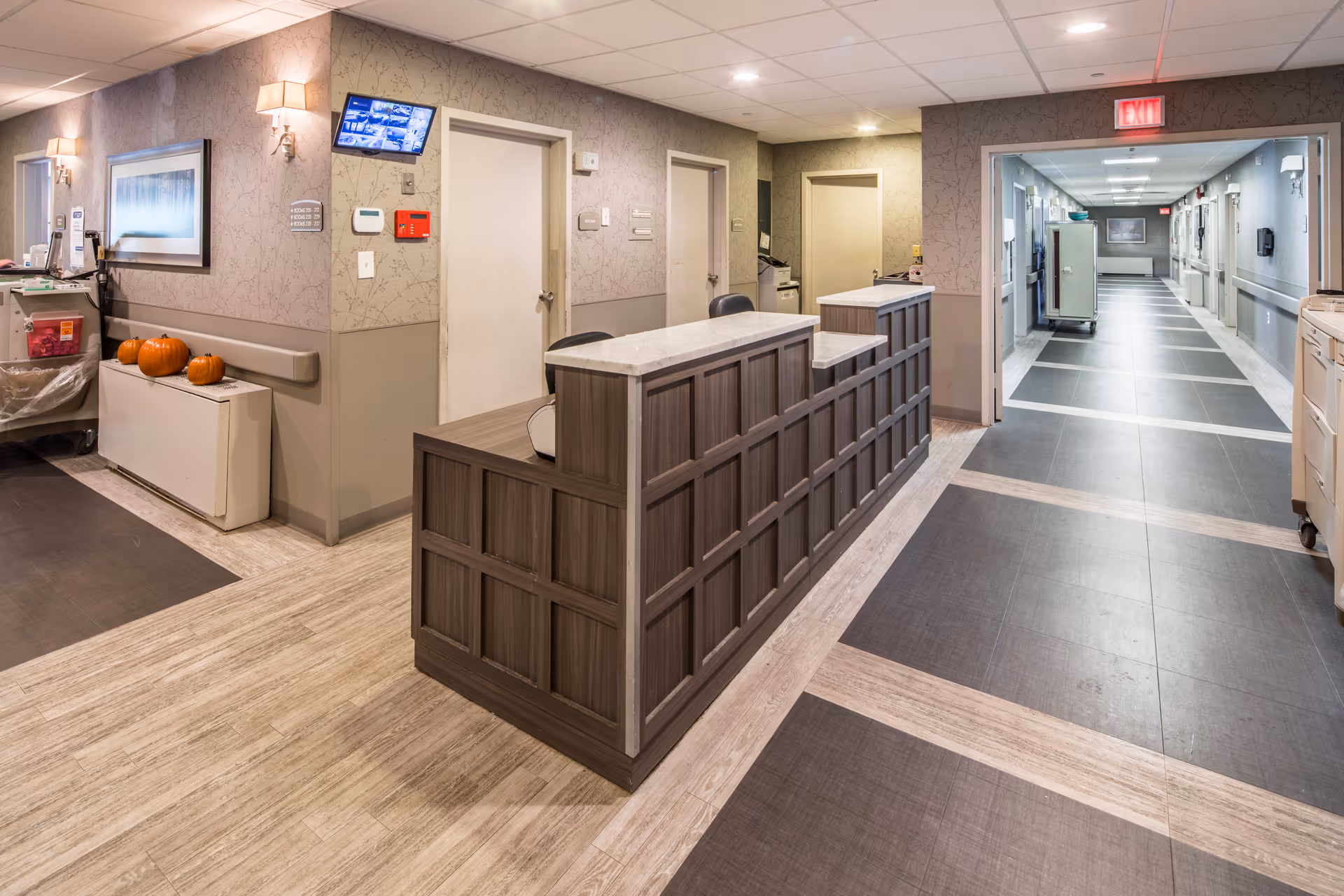 Reception desk area inside Highland Rehabilitation & Nursing Center with a wooden counter, chairs, and a hallway leading to patient rooms. The walls are decorated with light wallpaper and sconces, and there are pumpkins placed on a cabinet near the wall.