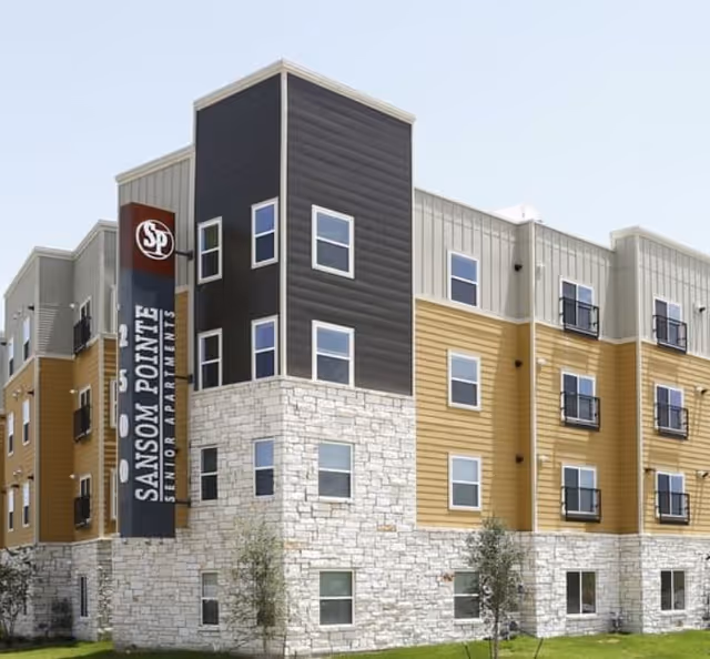 Exterior view of Sansom Pointe Senior Apartments, a modern multi-story building with a combination of stone, yellow siding, and dark paneling. The building has multiple windows and a large vertical sign displaying the name and logo.