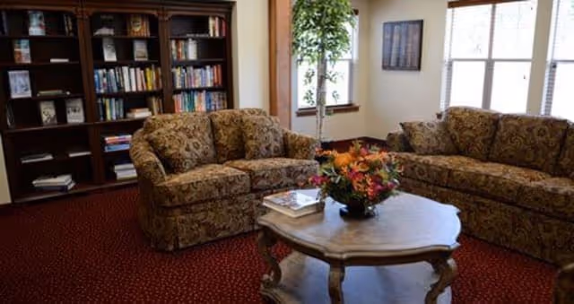 A cozy living room with two patterned sofas facing each other, a wooden coffee table with a floral centerpiece and books, a tall bookshelf filled with books, a potted plant near the window, and framed artwork on the wall.