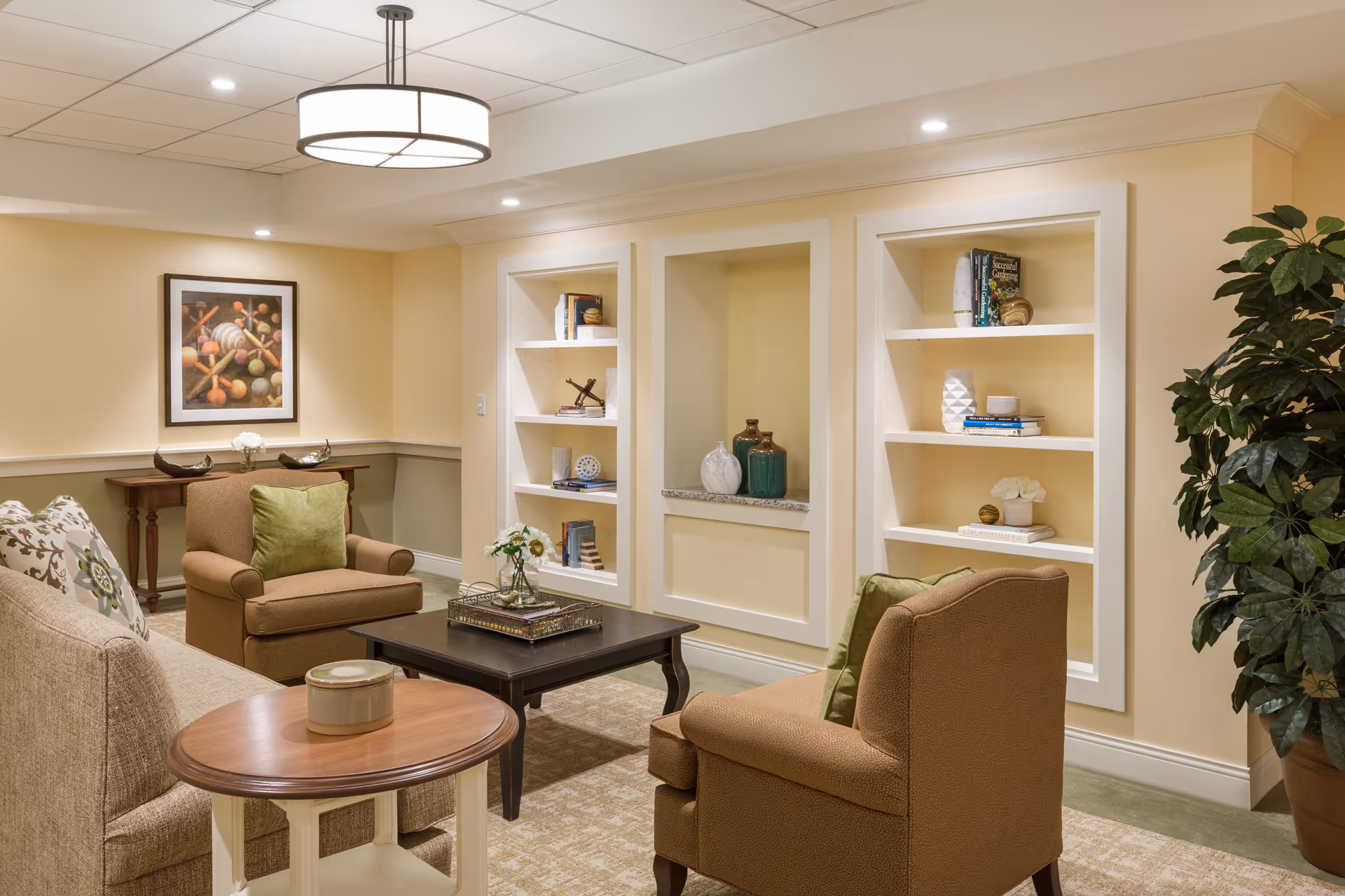 A cozy living room area with beige walls and carpet, featuring a sofa with patterned cushions, two brown armchairs with green pillows, a dark wooden coffee table with a decorative tray and flowers, built-in white shelves with books and decorative items, a framed artwork on the wall, a round side table, and a large potted plant in the corner. The ceiling has recessed lighting and a modern circular pendant light.
