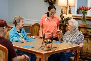 Three elderly individuals sitting around a table playing a bingo game with a staff member standing and smiling with them in a warmly decorated room with flowers and lamps in the background.