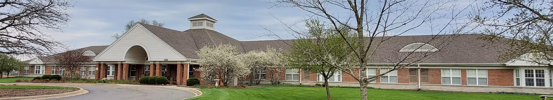 Wide exterior view of a single-story senior living facility building with a covered entrance, brick and siding facade, surrounded by green grass and trees with light foliage under a cloudy sky.