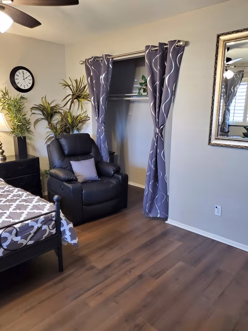 A cozy bedroom corner featuring a black recliner chair with a small pillow, a bed with patterned bedding, a nightstand with a lamp, several potted plants, a wall clock, a large mirror, and a closet with curtains instead of doors. The room has wooden flooring and neutral-colored walls.